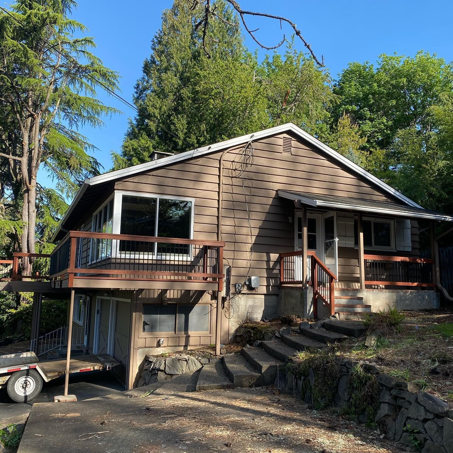 Brown cabin with a front deck and stairs, surrounded by trees on a wooded hillside.