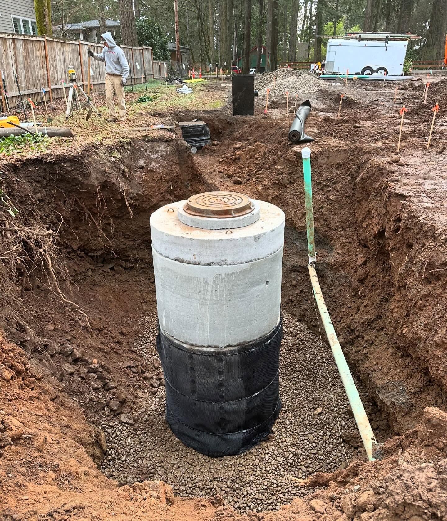 A concrete septic tank stands in a dug pit at a wooded construction site.