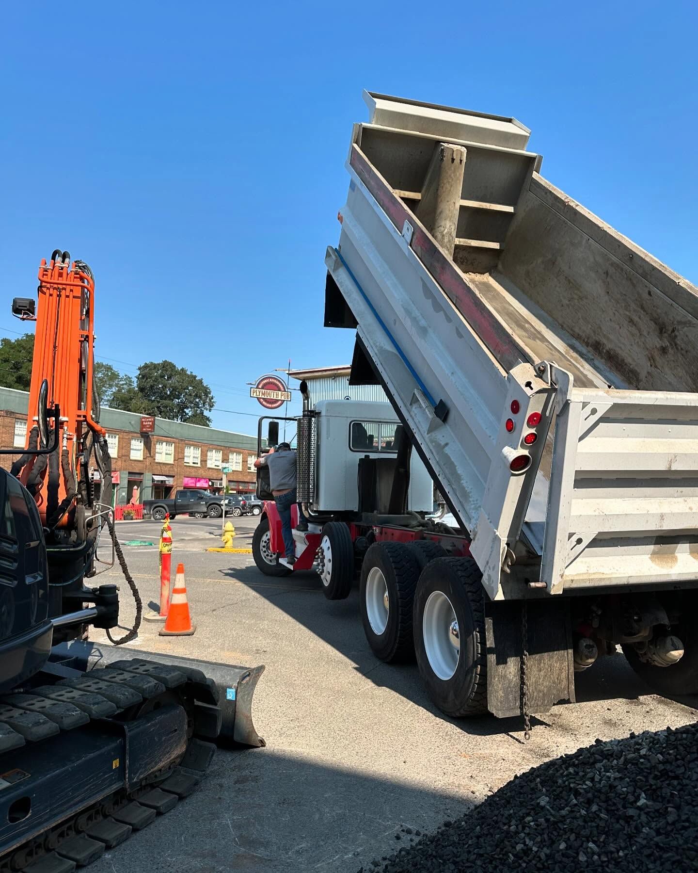 Dump truck raising its bed at a roadwork site, with an excavator and traffic cones nearby.