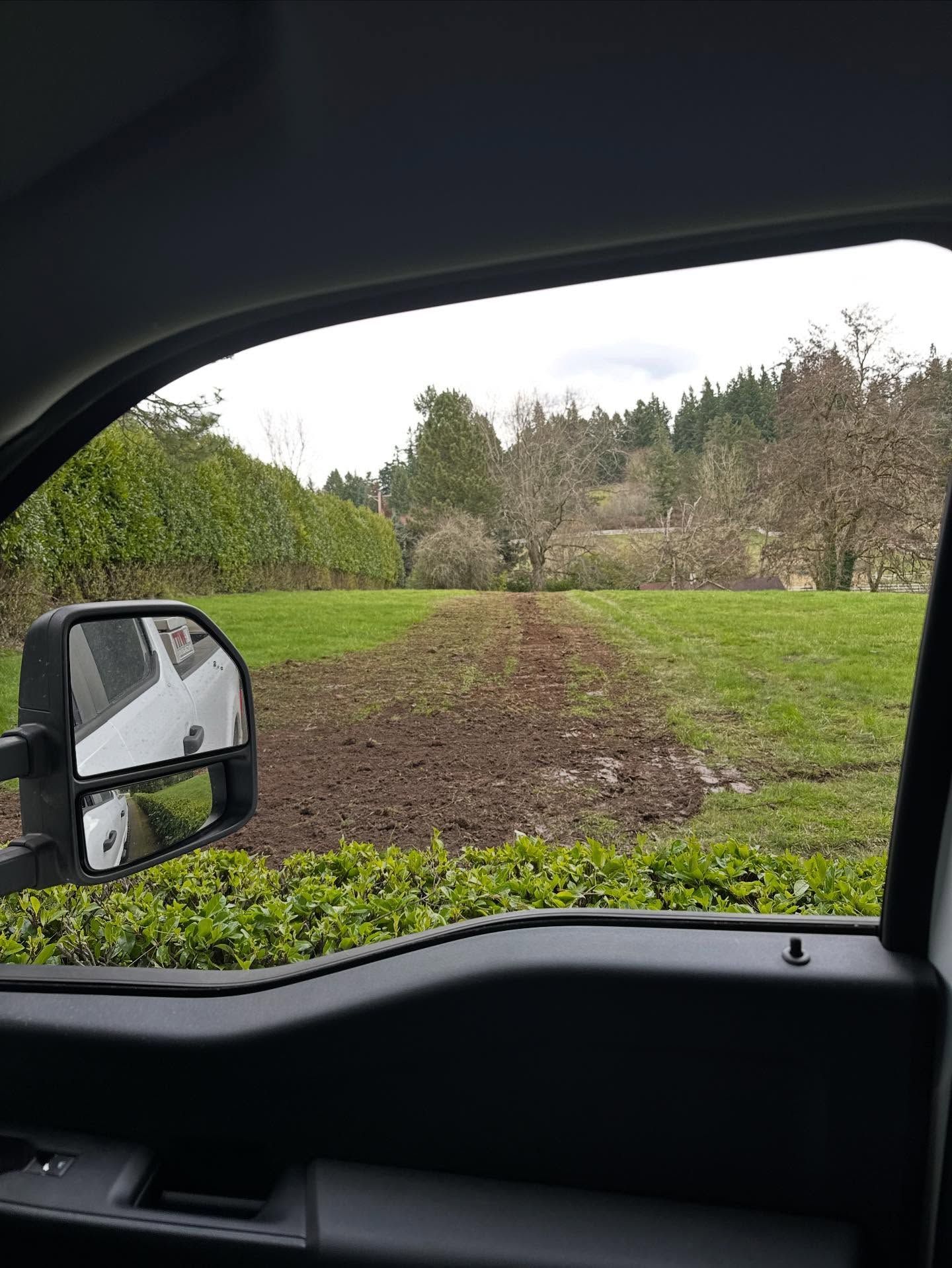 View from a vehicle of a muddy patch in a green field with trees in the background.