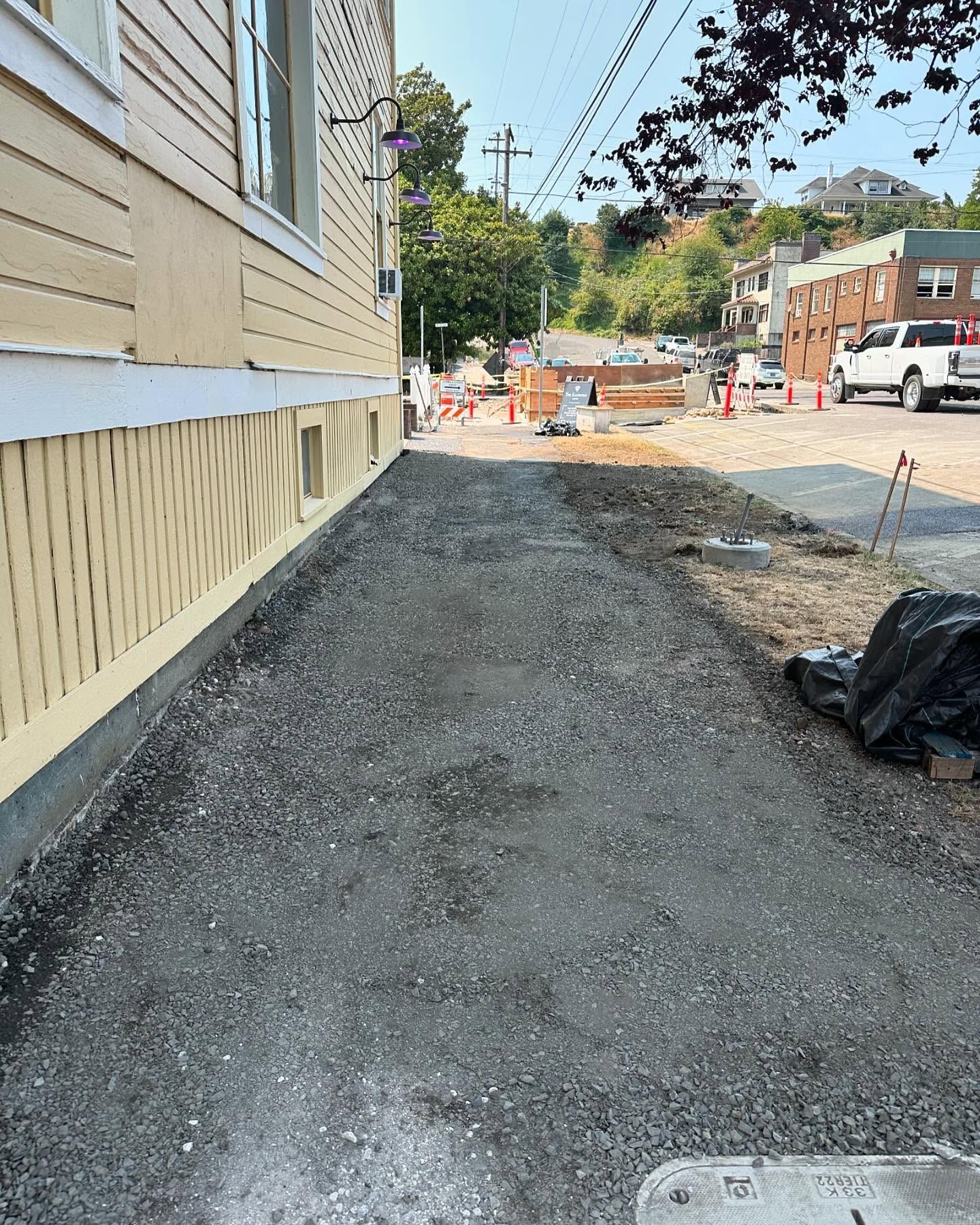Sidewalk beside beige building under construction, with gravel path, orange cones, and parked trucks ahead.