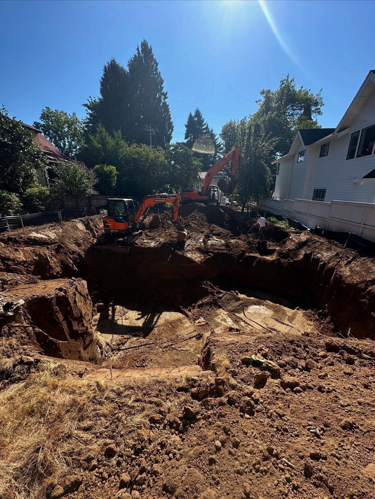 Excavator digging a large pit in a sunny backyard beside a white house and trees