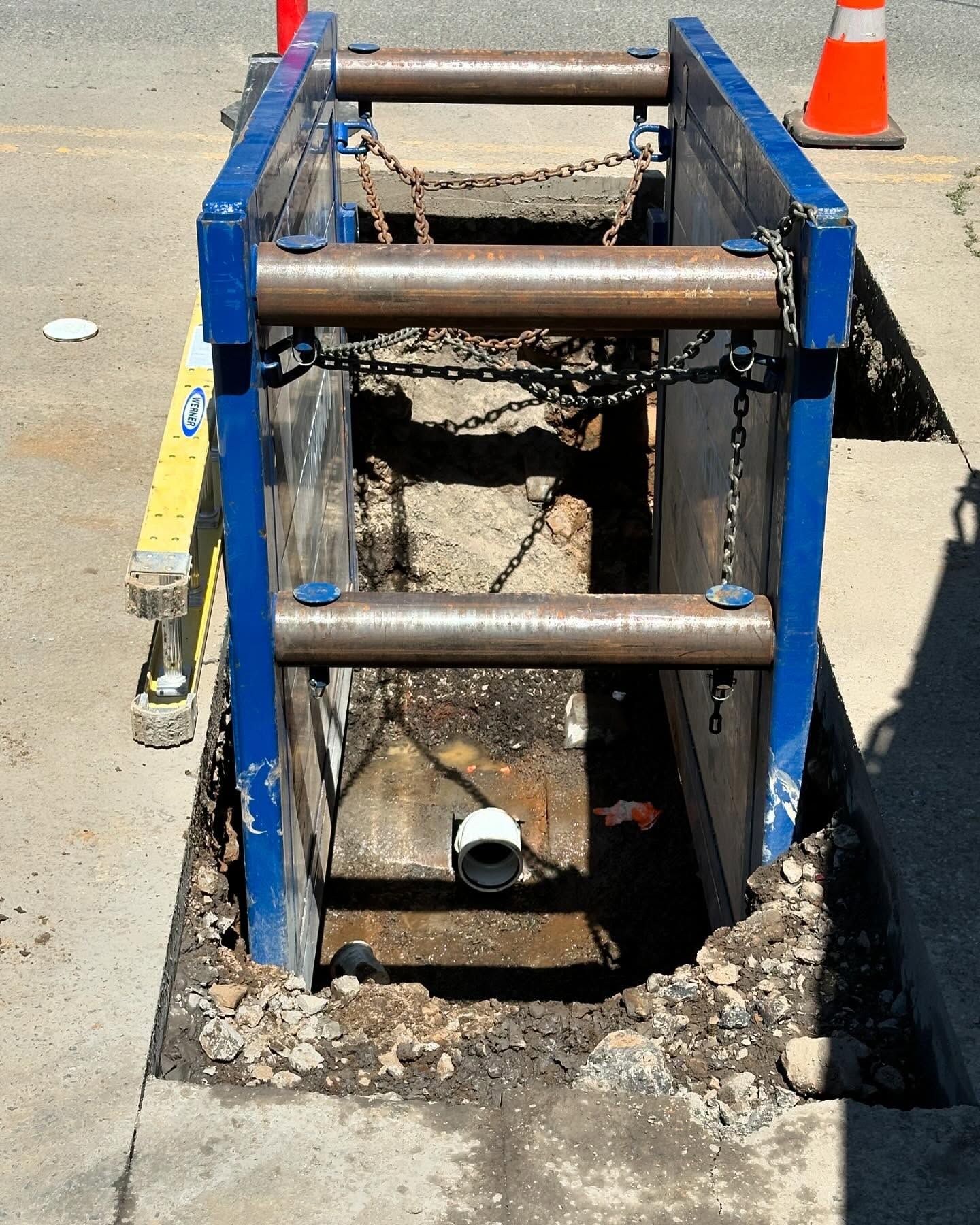 Blue trench shoring frame around an open utility excavation with an orange cone nearby.
