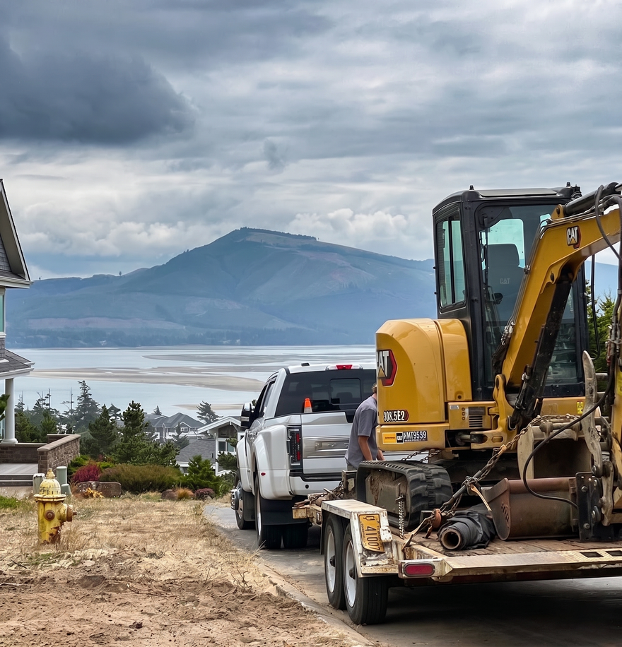 Yellow excavator on a flatbed trailer beside a white truck, overlooking water and mountains under cloudy skies