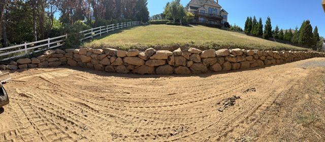 Freshly graded dirt slope with a stacked stone retaining wall and house on a hill above