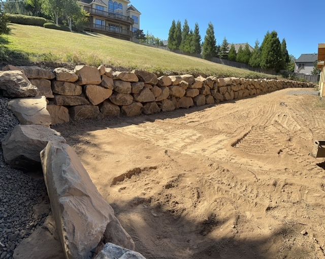 Sandy construction area bordered by a stone retaining wall beside a grassy slope and homes