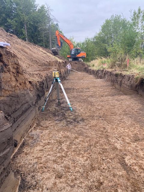 Survey tripod on a dirt trench with an orange excavator in the background.