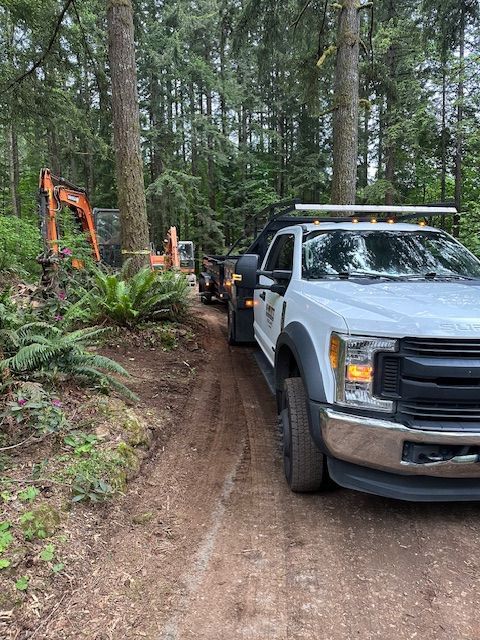 White utility truck on a muddy forest road beside an orange excavator.
