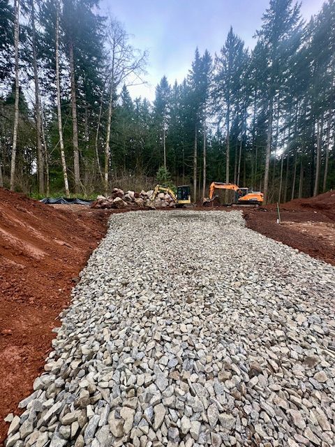 Gravel road base being built in a forest clearing with excavator and stacked logs in the background