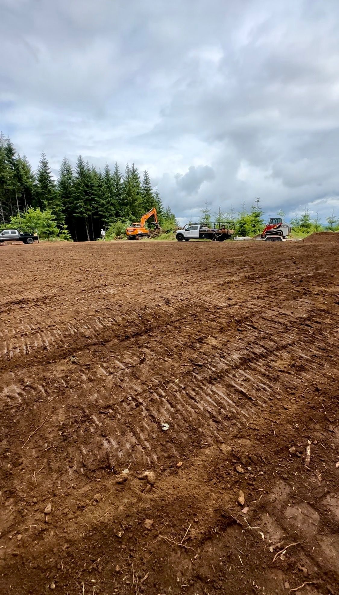 Freshly graded dirt lot with construction equipment, evergreen trees, and cloudy sky in the background