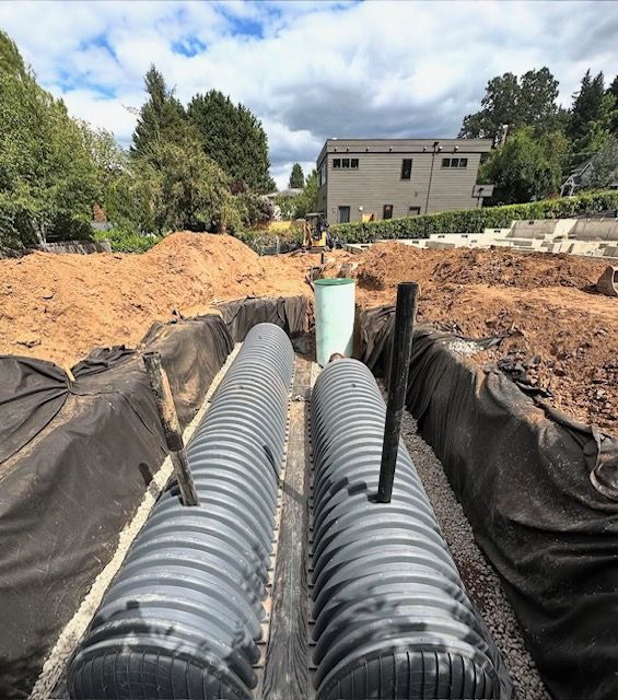 Construction trench with two large corrugated drainage pipes and black liner, dirt piles, and houses in background