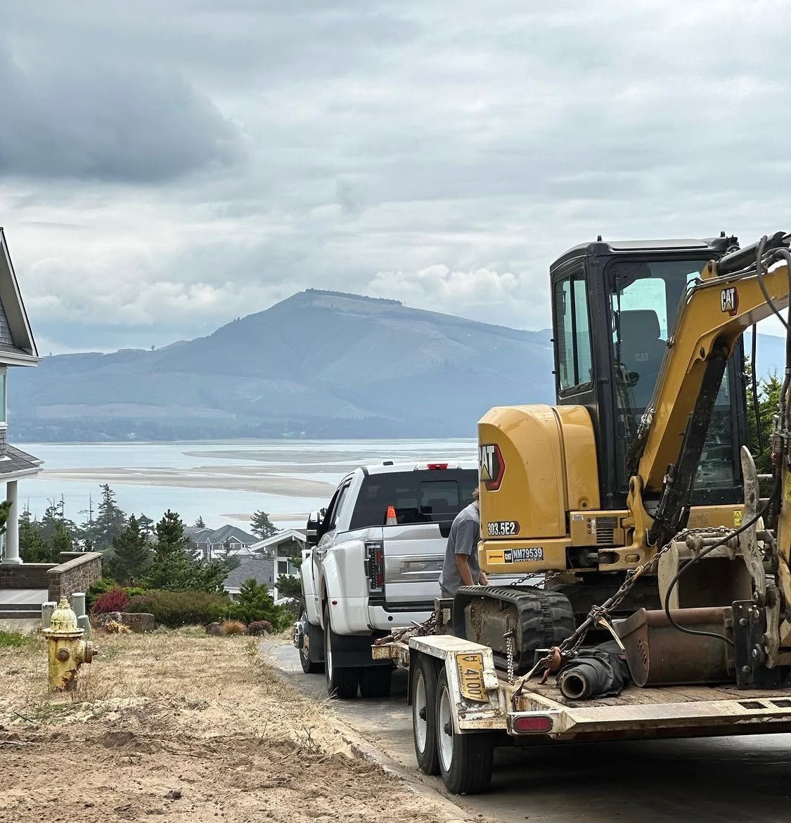 Yellow excavator on a trailer in a dirt driveway overlooking a bay and mountain under cloudy skies