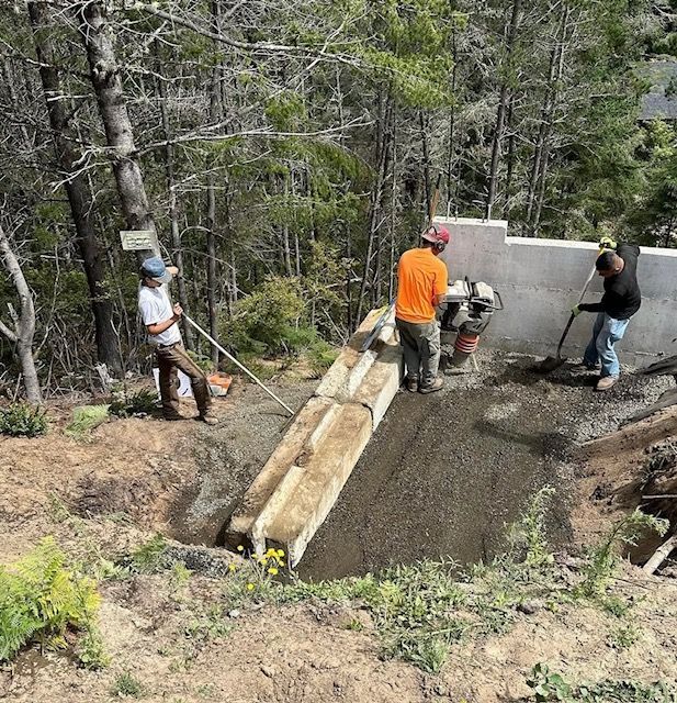 Workers pouring concrete for a small bridge or retaining wall in a forested area