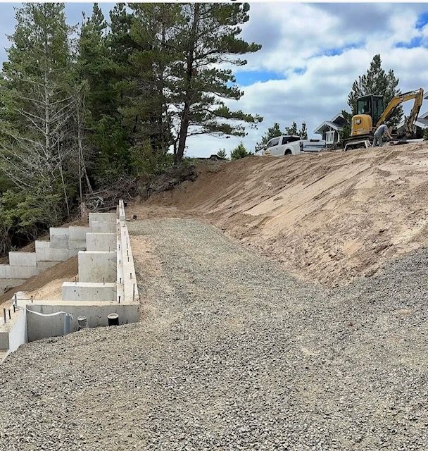Construction site with gravel road, retaining wall, excavator, and pine trees on a hillside