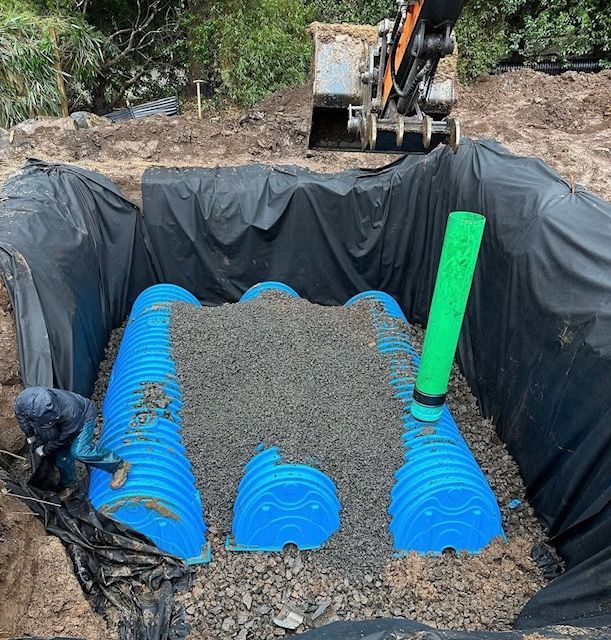 Excavation pit lined with black tarp, blue tanks, gravel fill, and a green pipe under construction