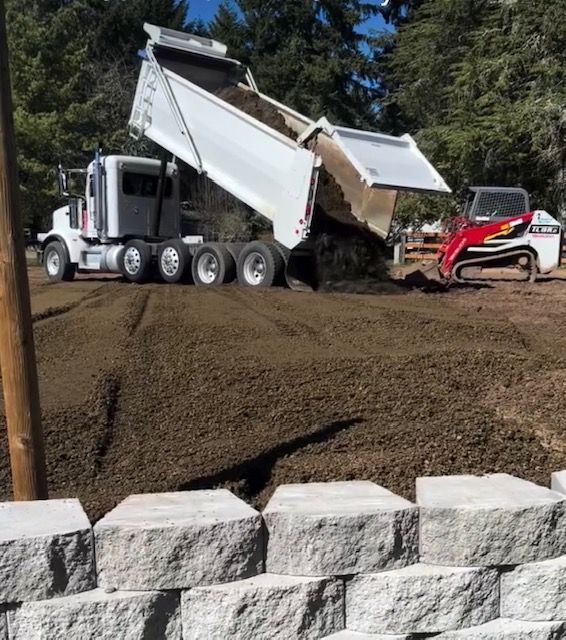 White dump truck unloading dirt on a construction site beside a red skid steer, with stone blocks in front.