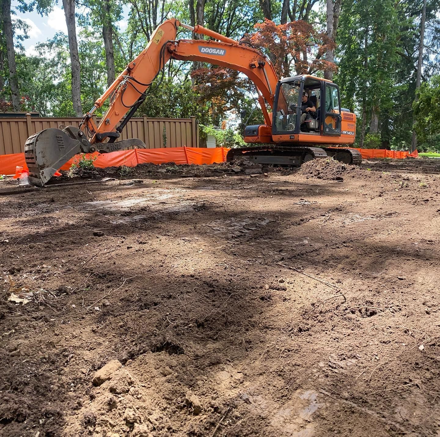 Orange excavator leveling dirt in a wooded construction area, with orange safety fencing nearby.