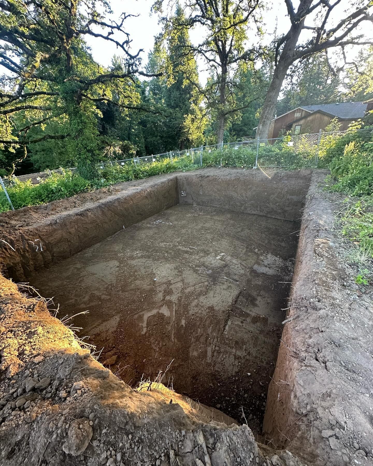 Rectangular excavated pit in a wooded garden with dirt walls and floor