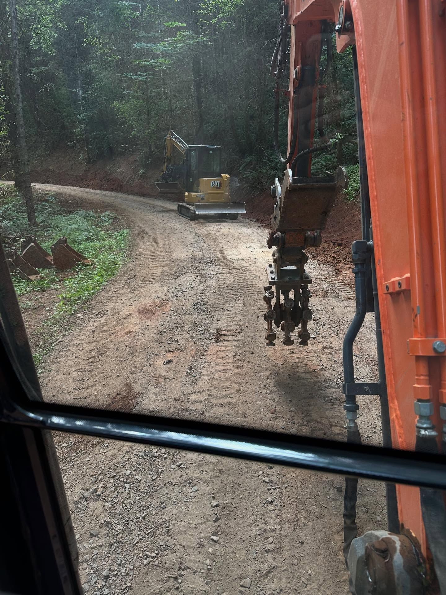 Excavator arm with claw on a muddy forest road, viewed from inside another excavator cab.