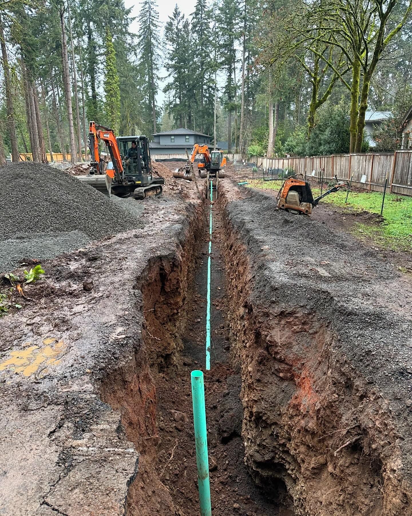 Excavator digging a deep trench with green pipe laid along it in a muddy forested worksite