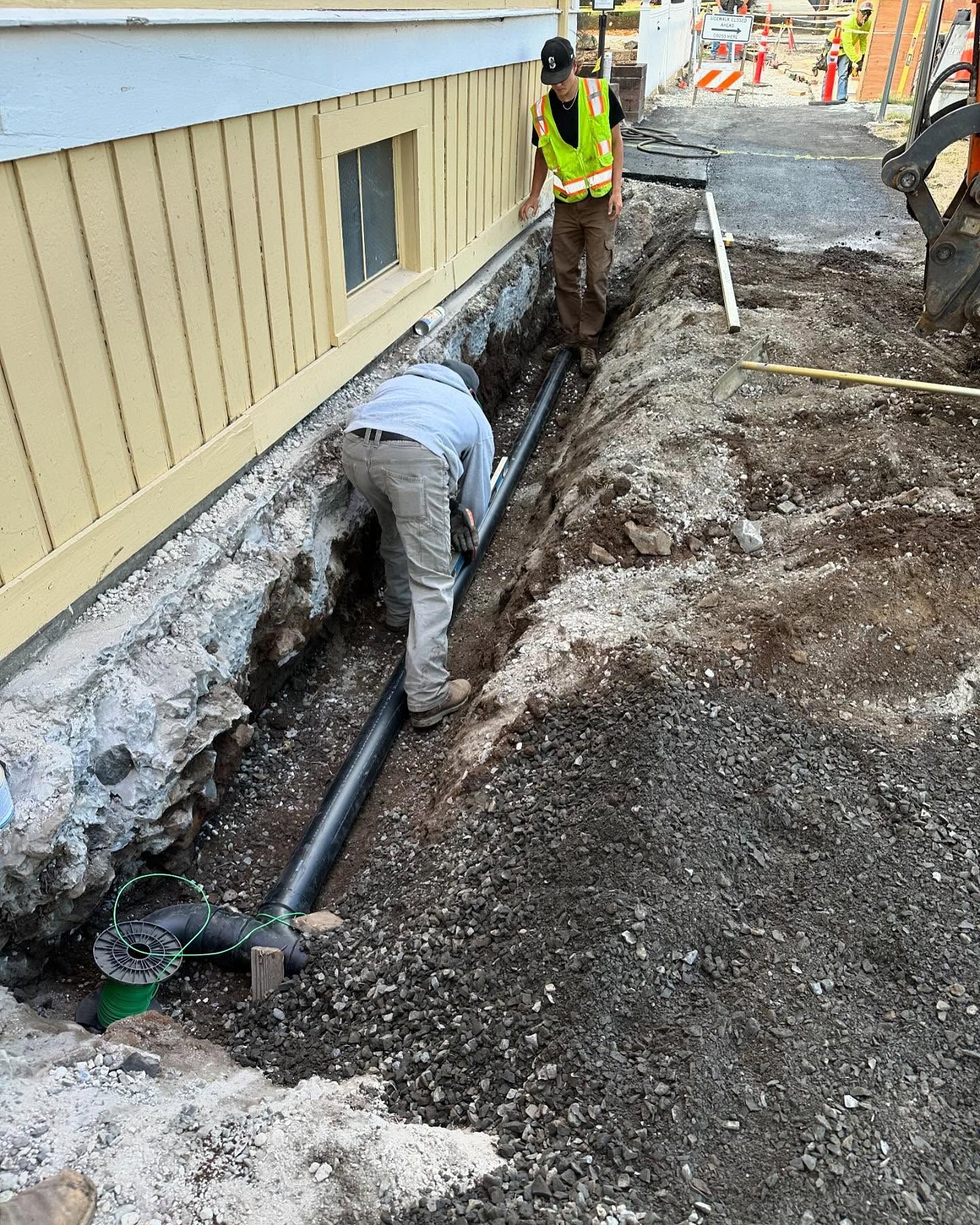 Workers digging a trench beside a beige building, exposing a black pipe along a muddy construction site.