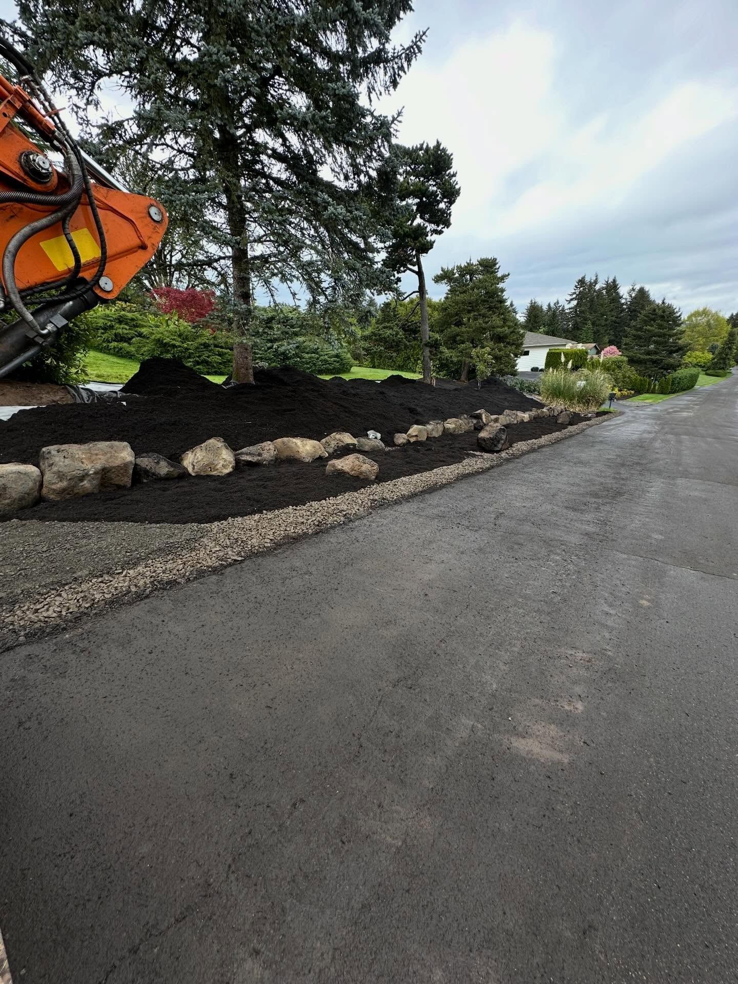 Roadside landscaping with rocks and mulch beside a freshly paved road, with an excavator at left.