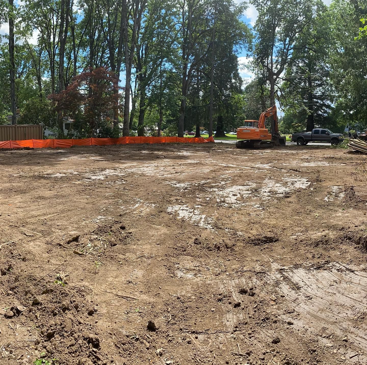 Cleared dirt lot with orange safety fencing, trees, and an excavator near a pickup truck in the background