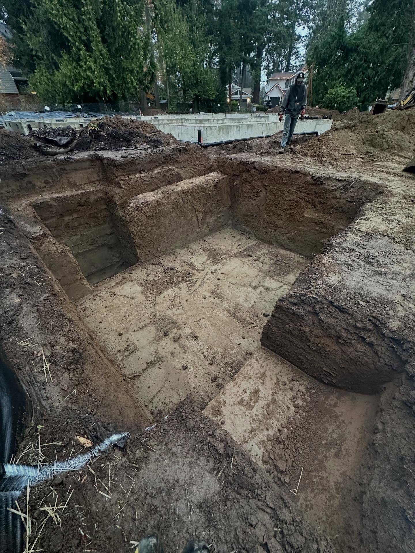 Large rectangular excavation site with muddy soil and a worker in the background near trees