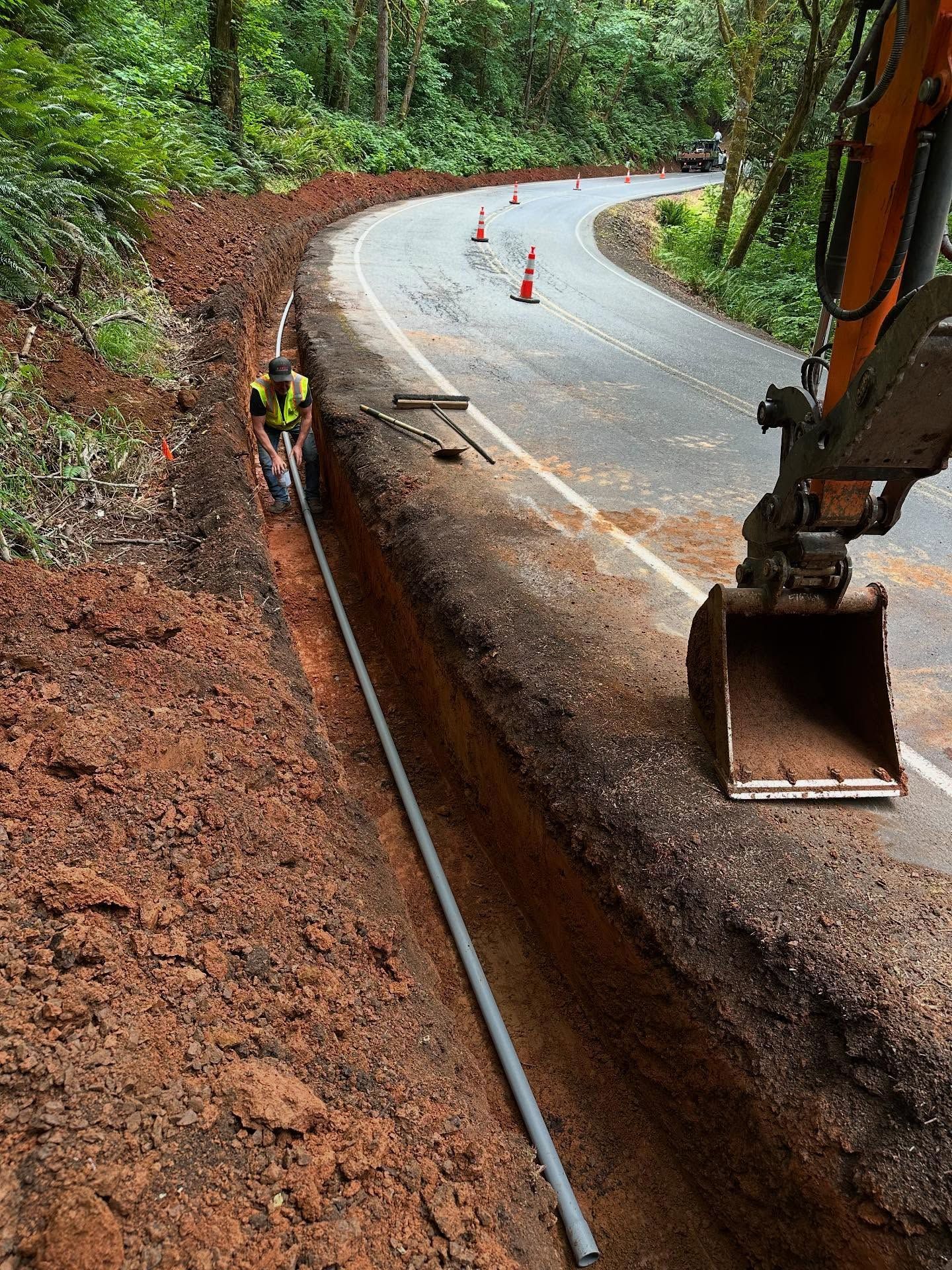 Road construction beside a forested road, with an excavator digging a trench and orange cones ahead.