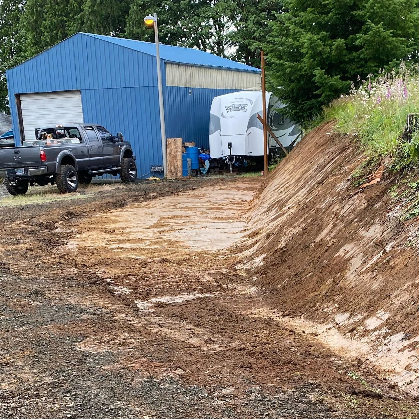Blue shed beside muddy dirt driveway with parked truck and white trailer near a sloped embankment