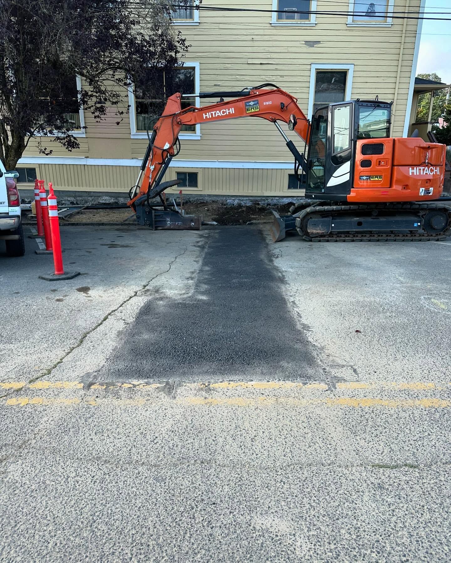 Orange excavator digging near a yellow house beside a street with traffic cones and a closed road