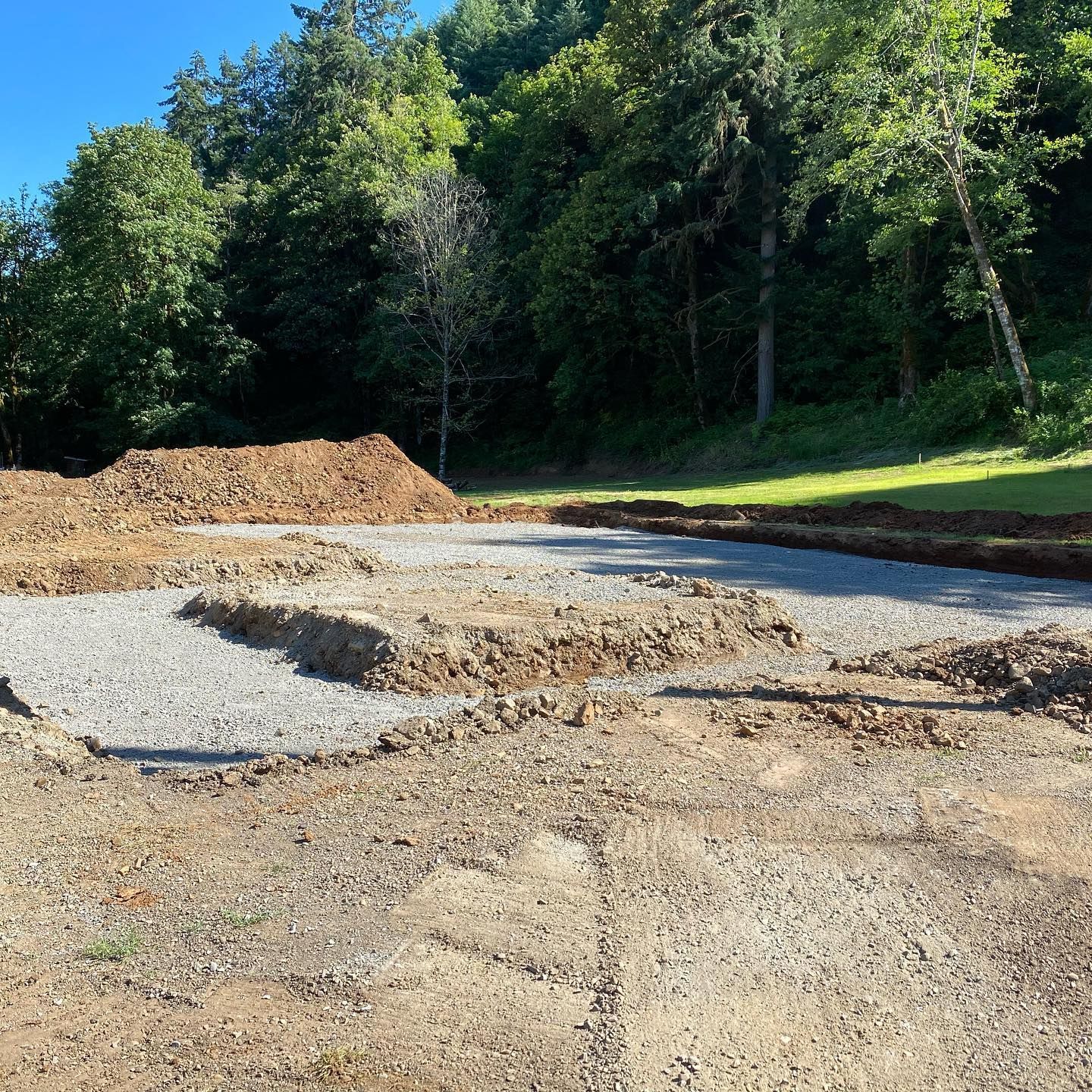 Gravel and dirt construction site bordered by dense green trees under a bright blue sky
