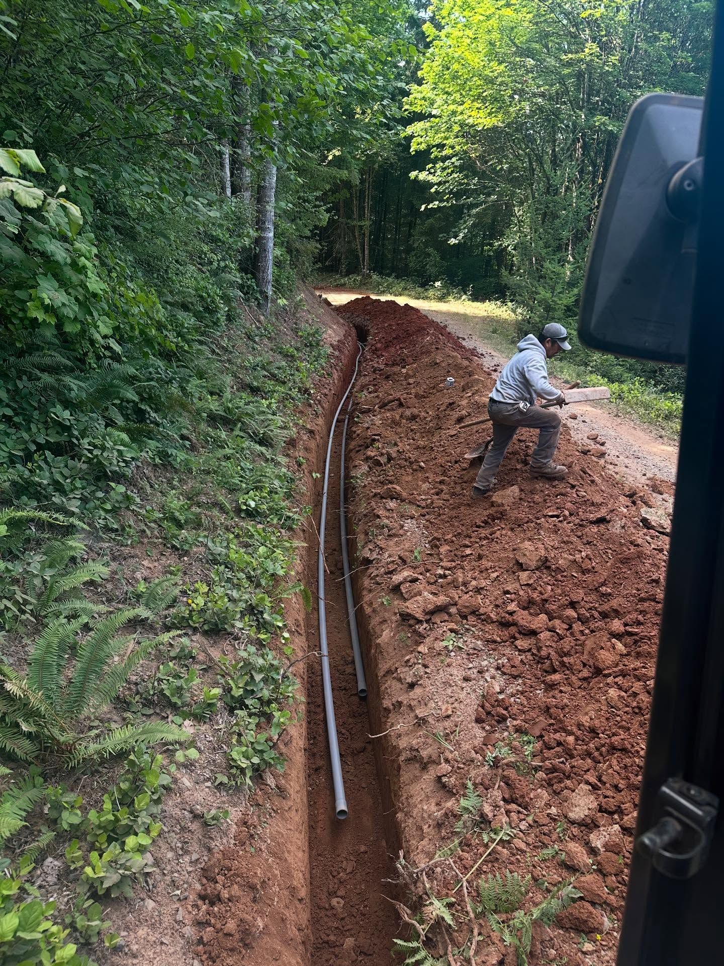 Worker digging a trench along a forested dirt road beside a long black pipe