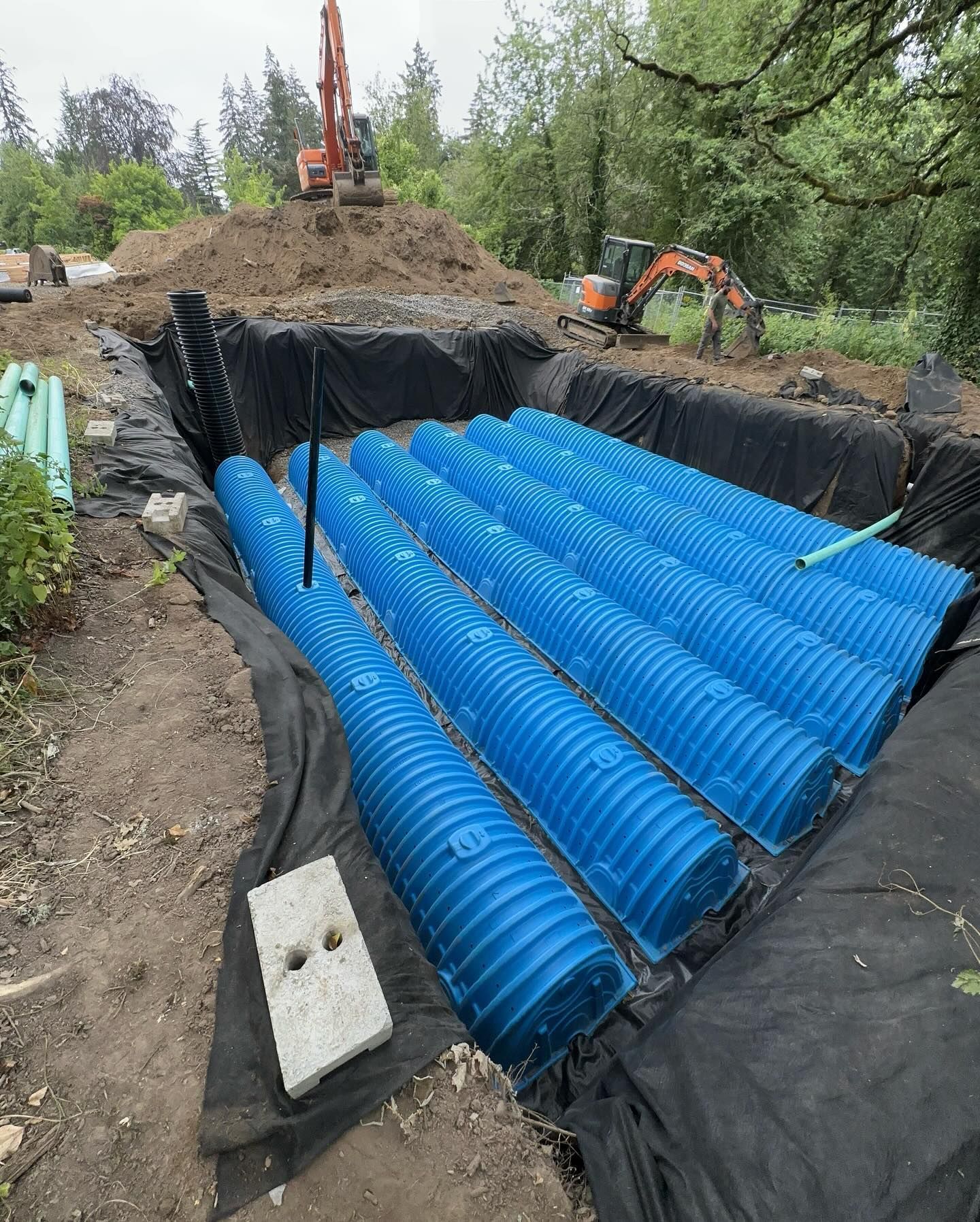 Construction trench lined with black tarp and blue corrugated pipes, with an excavator in the background.