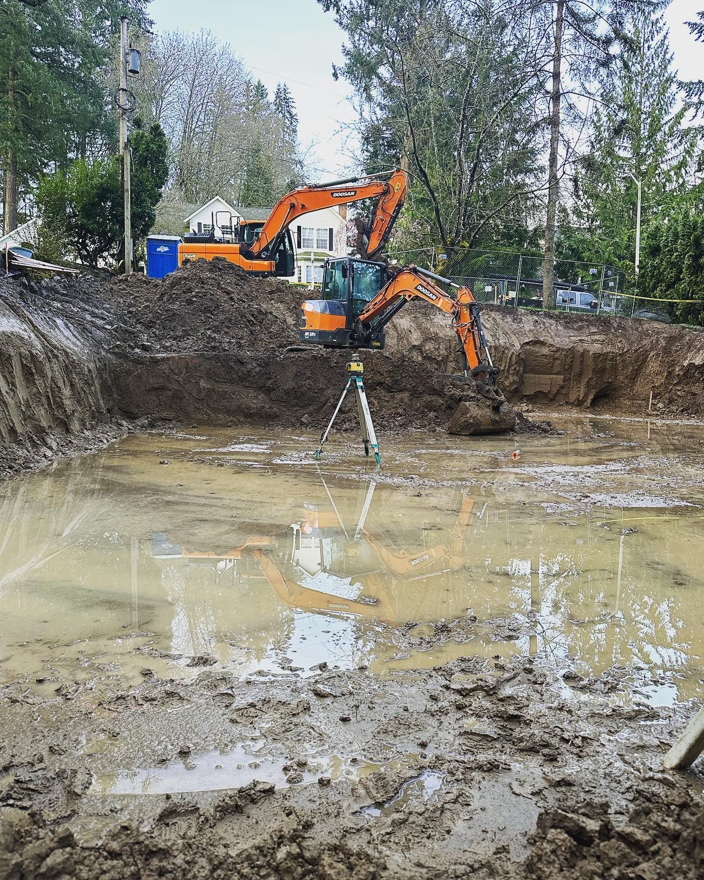 Orange excavator working in a muddy excavation pit beside standing water and trees