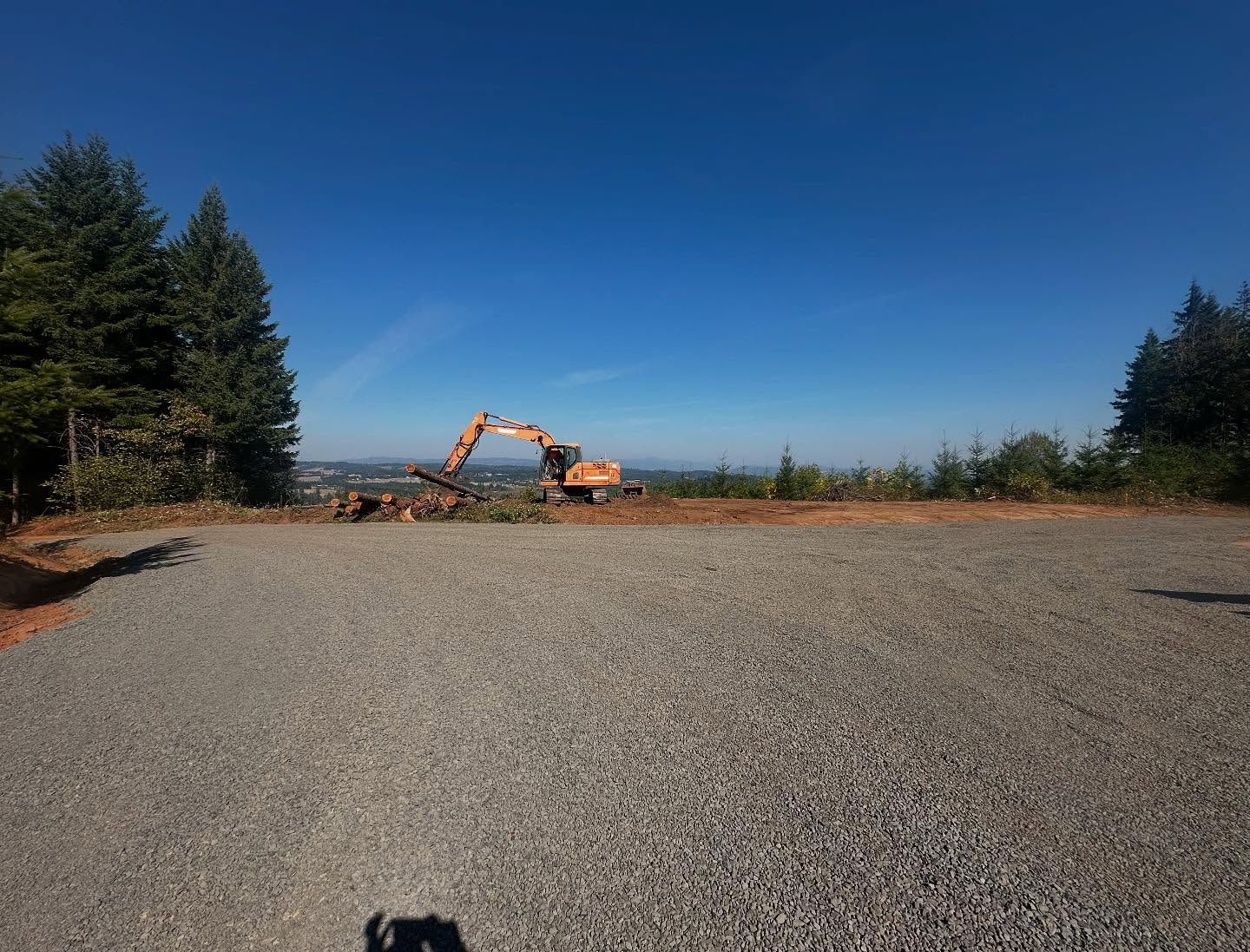 Excavator working on a gravel clearing with trees under a clear blue sky