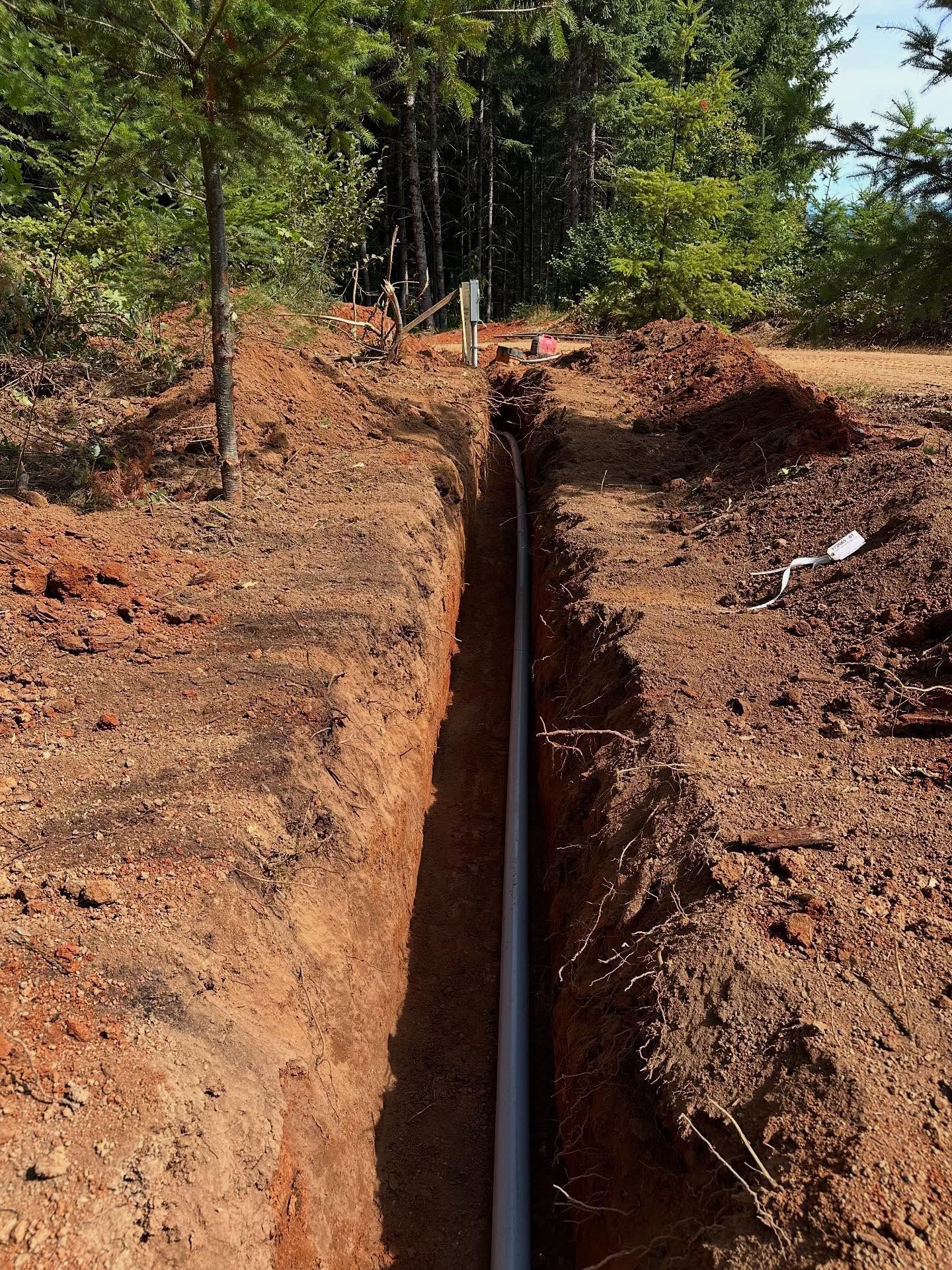 Narrow trench cut into red soil with a white pipe running along the bottom in a wooded area