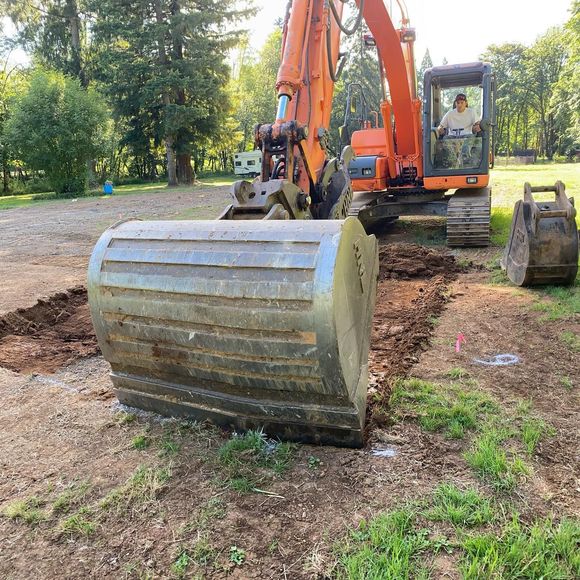 Orange excavator with a large bucket in a grassy dirt lot, surrounded by trees.