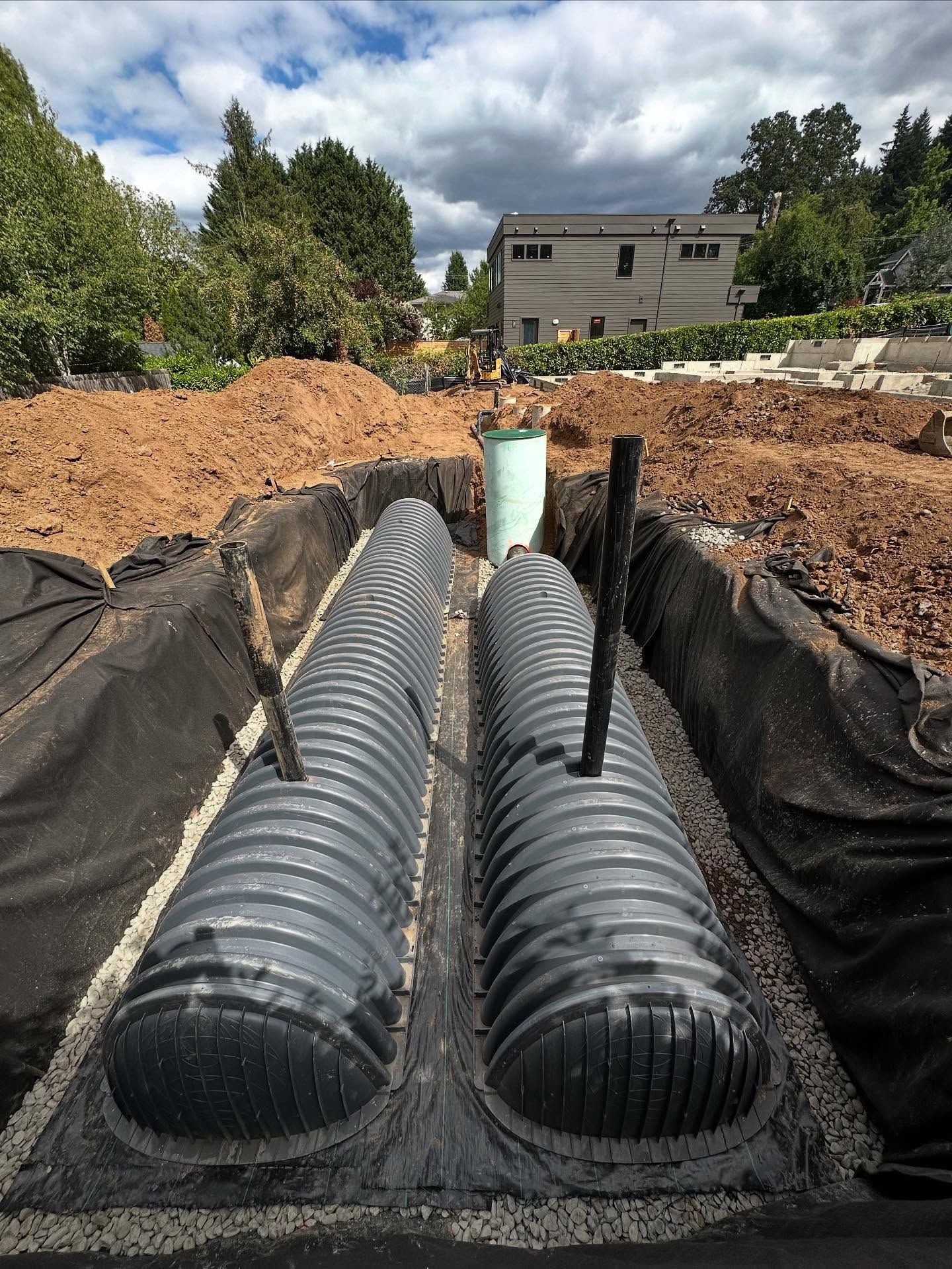 Two black corrugated drainage pipes installed in a trench at a construction site with dirt piles and trees nearby