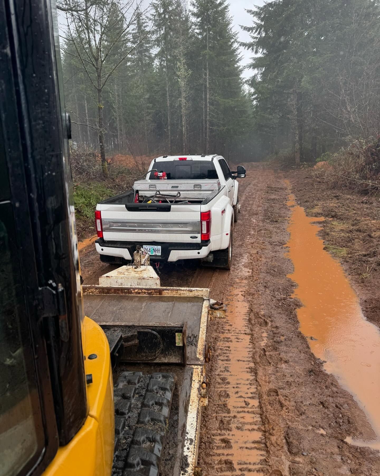 White pickup truck on muddy forest road beside an excavator, with rain and puddles in the background