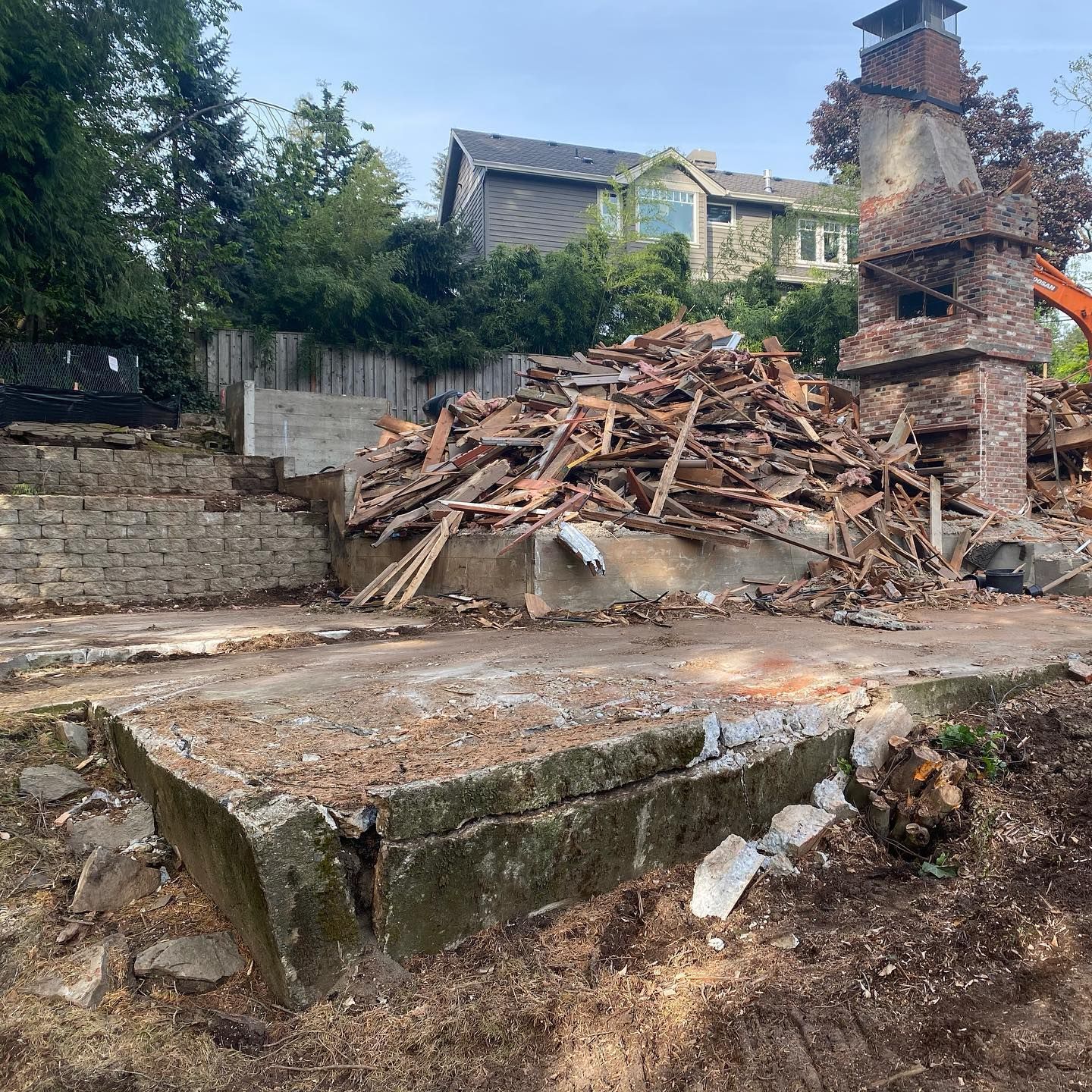 Collapsed stone wall and rubble pile in a residential yard, with a damaged chimney and houses behind.