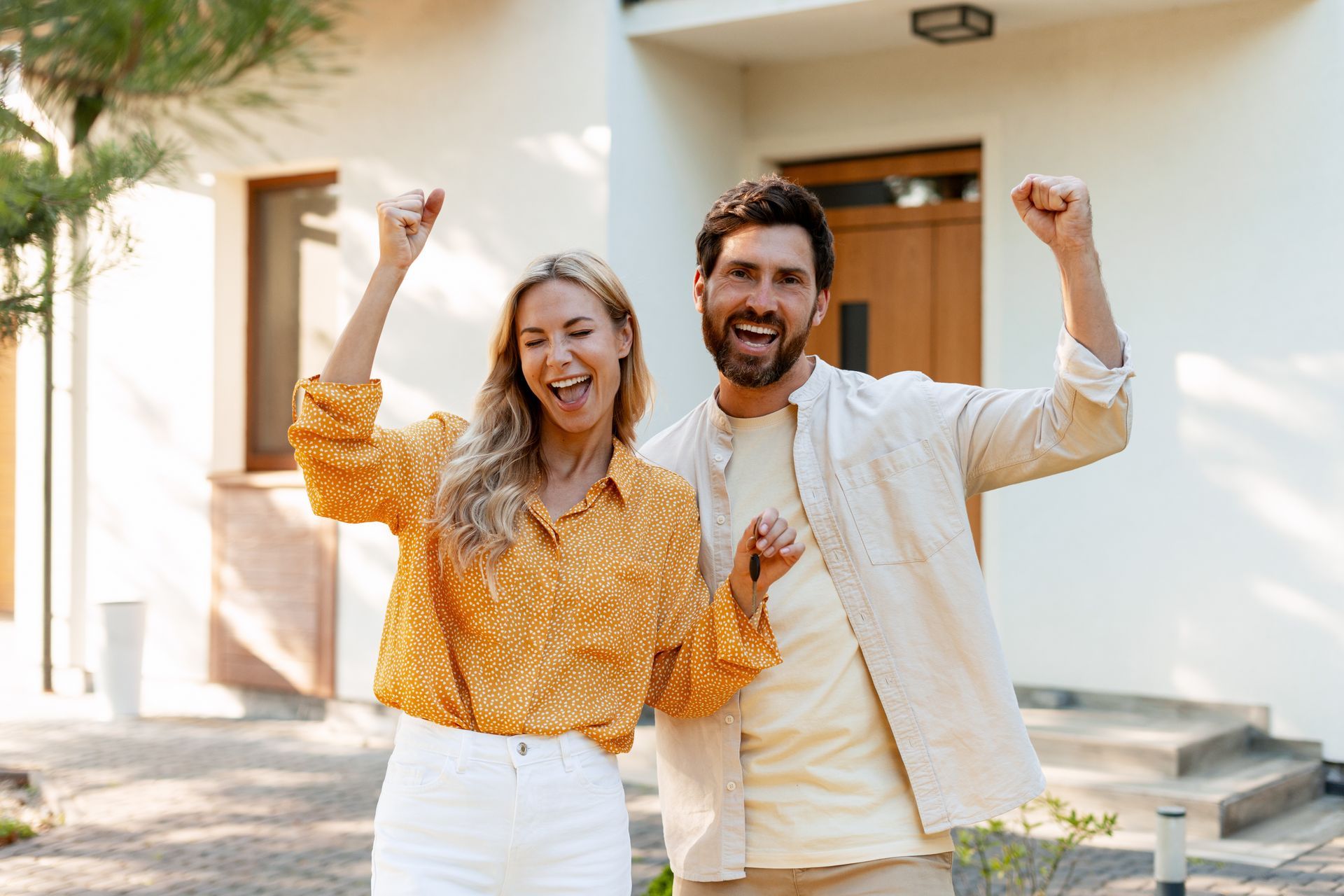 Two people cheering outside a house, smiling with raised fists, one in a yellow shirt and one in a white jacket