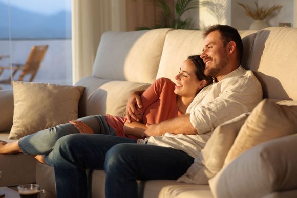 Couple relaxing together on a sofa in a cozy living room, smiling and cuddled under warm light