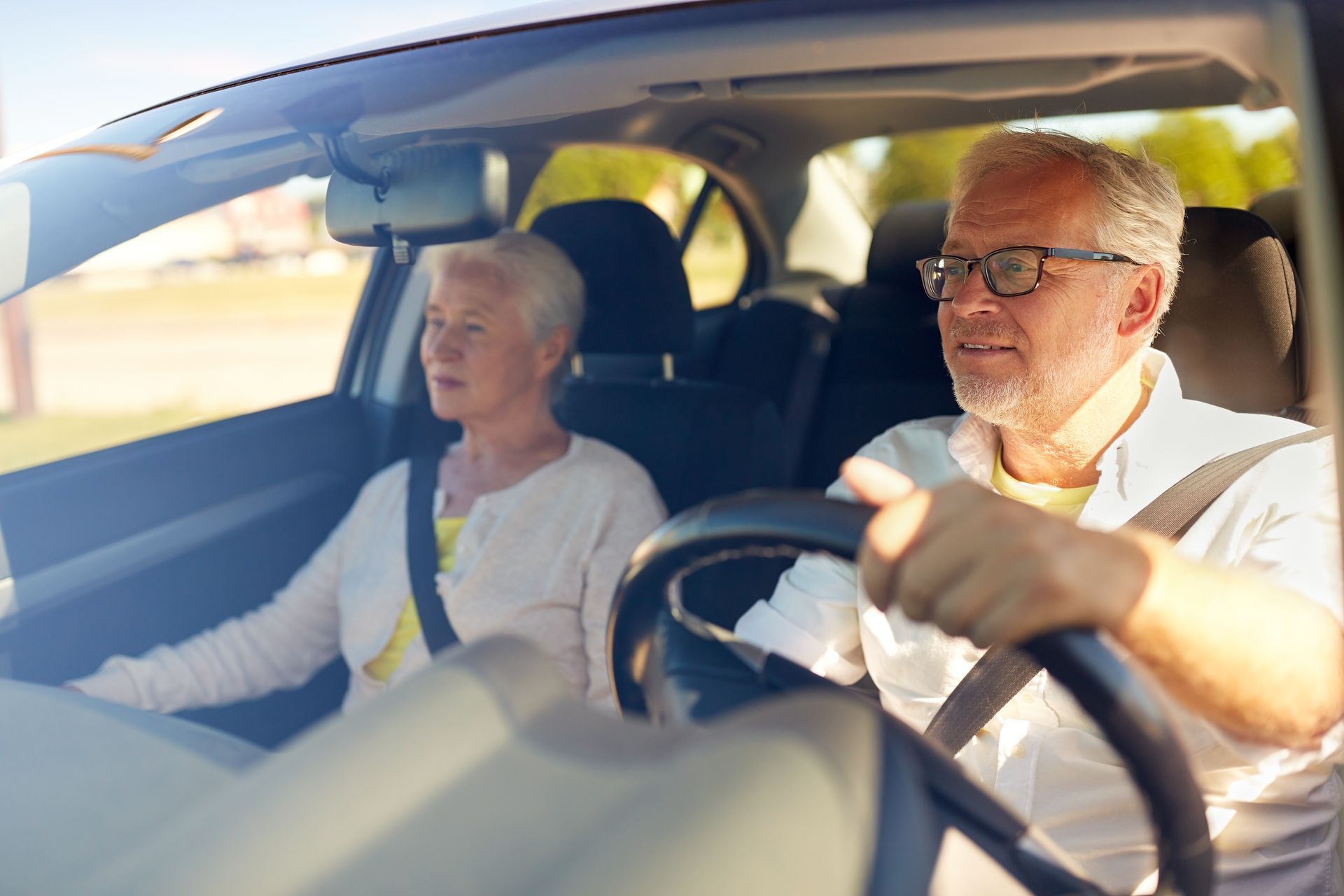 Two people driving in a car, with the driver focused on the road and bright sunlight through the windows.