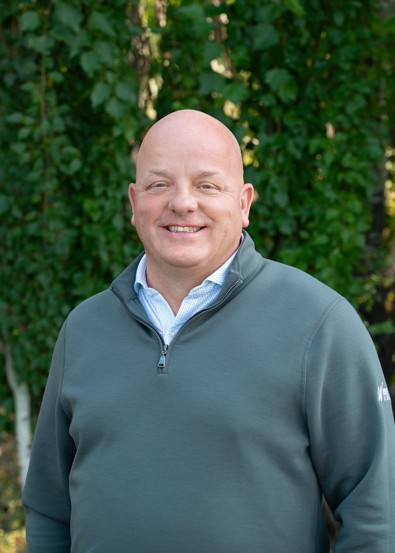 Someone wearing a grey quarter-zip sweater and a light-colored button-down shirt, smiling in front of a leafy green backdrop.