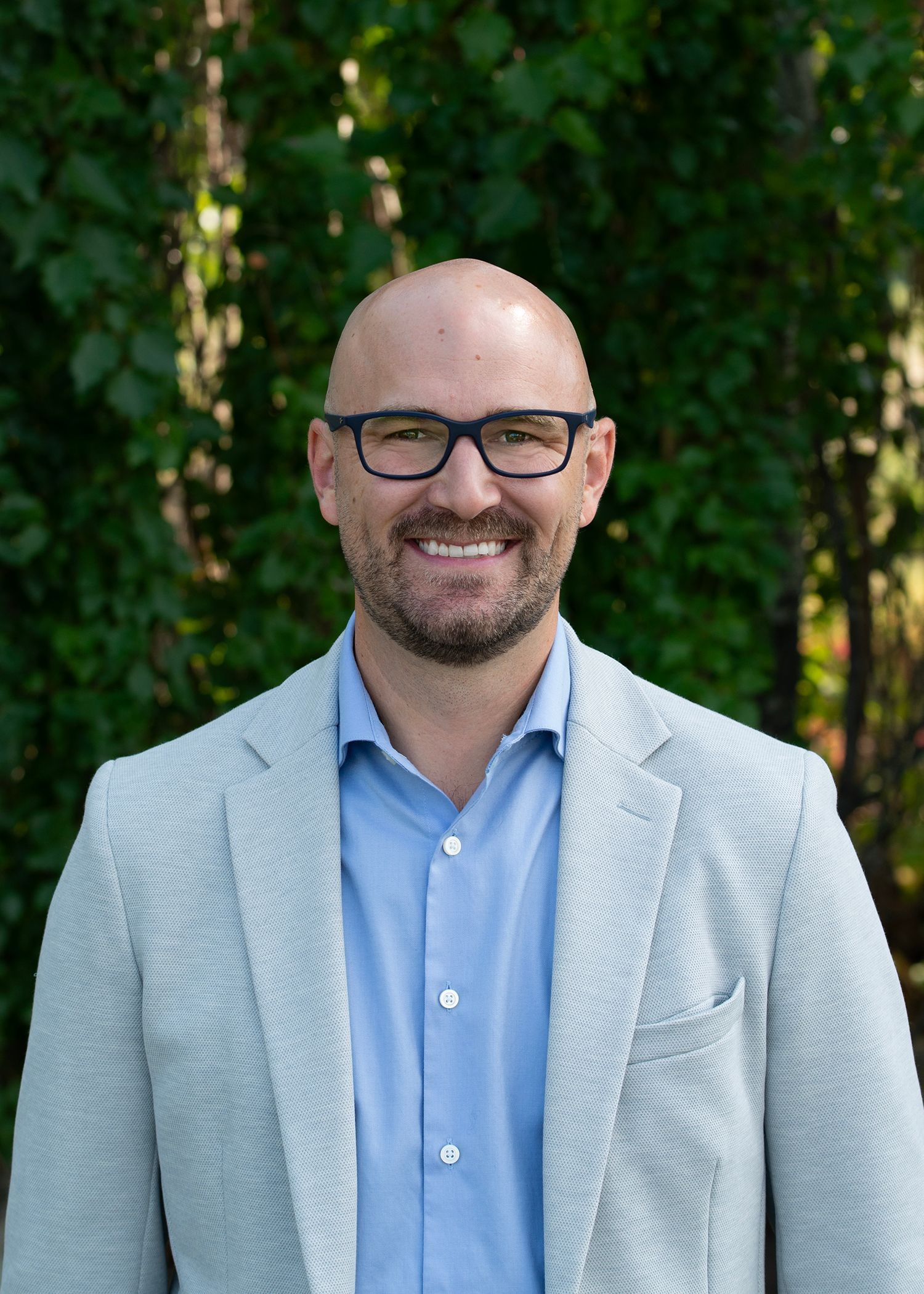 A person with a shaved head and glasses smiles, wearing a blue button-down shirt and a textured gray blazer outdoors.