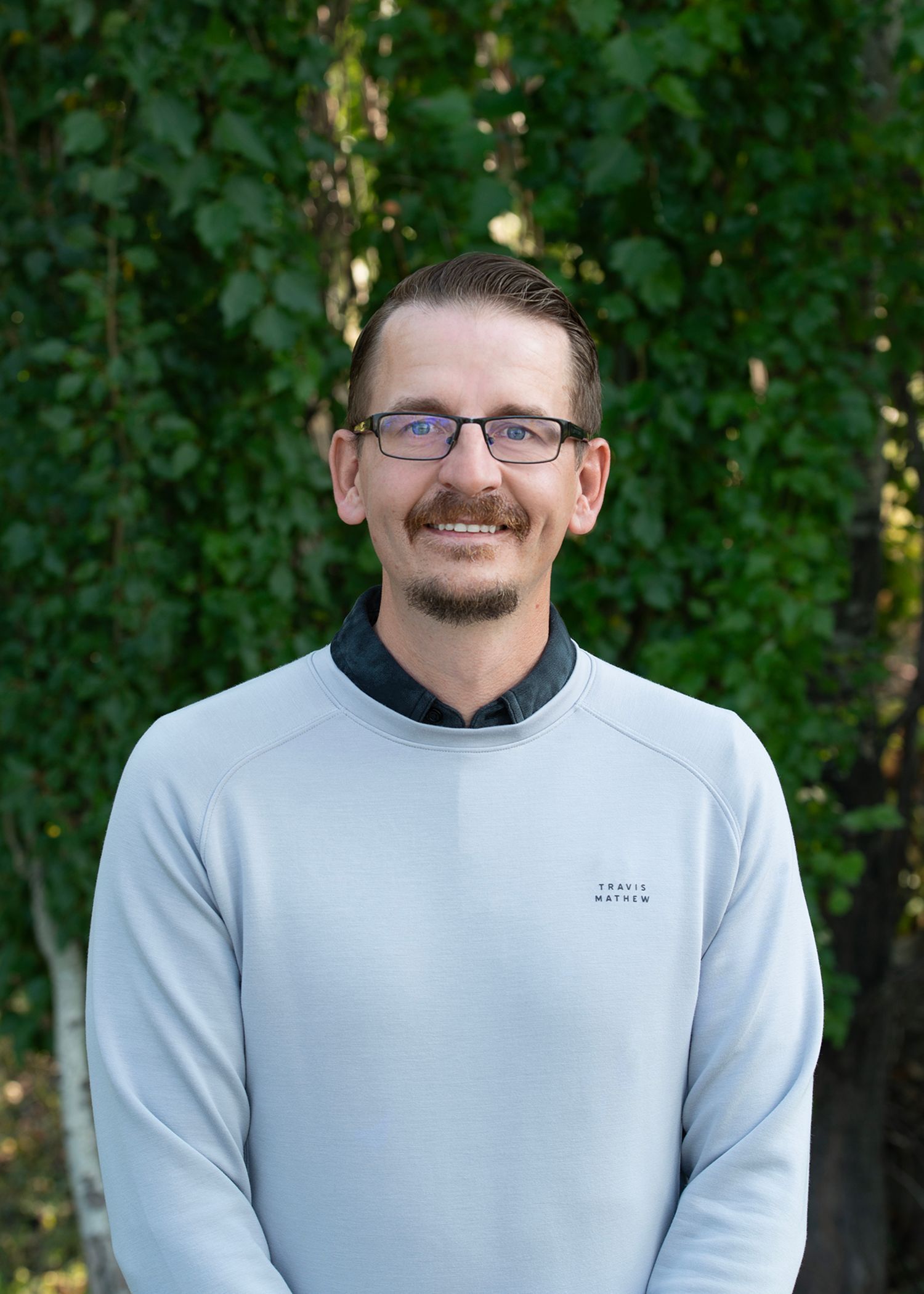 A smiling person with glasses and facial hair wears a light blue sweater, posed in front of a green leafy background.