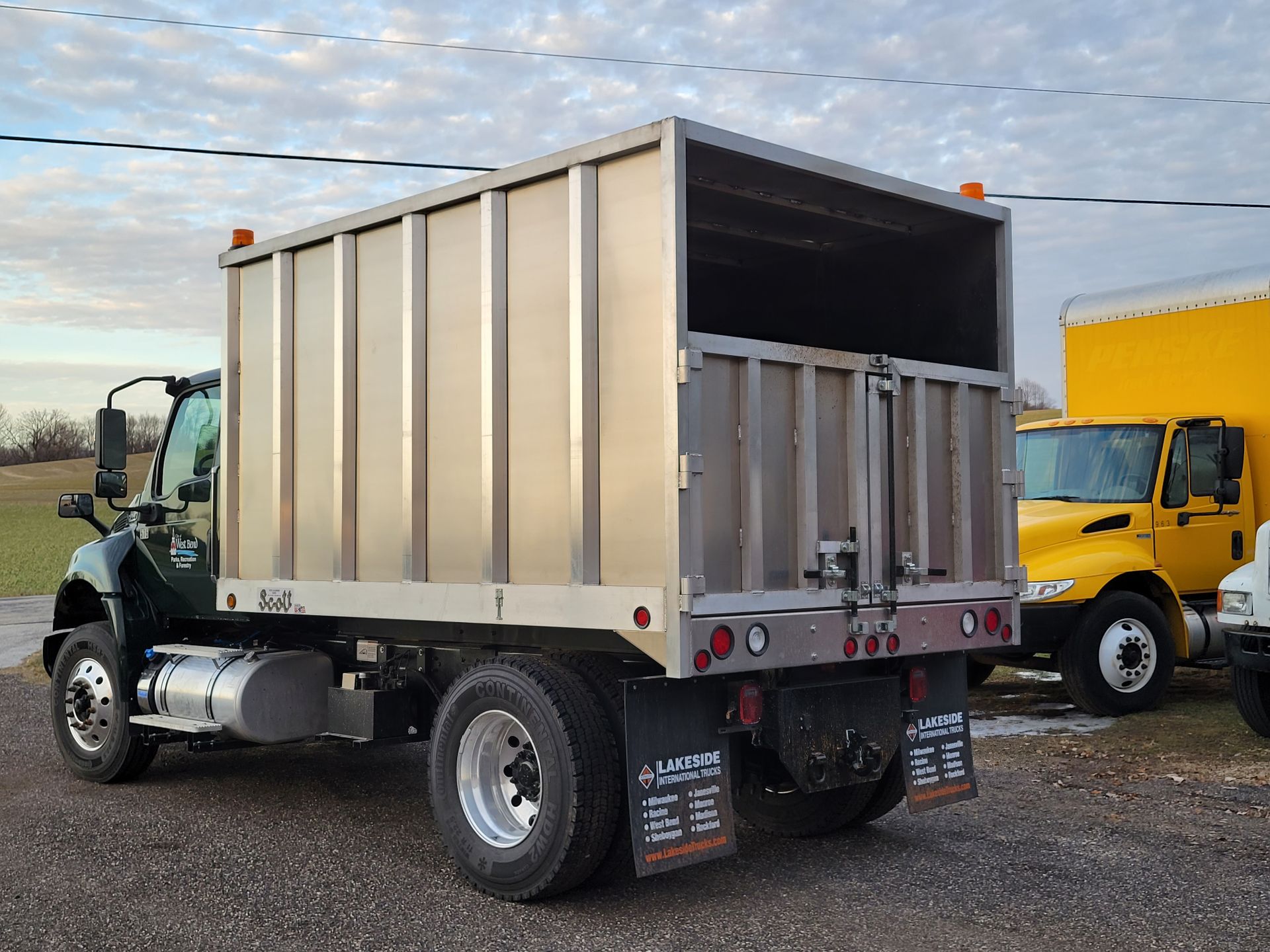 A truck with a stainless steel body is parked next to a yellow truck.