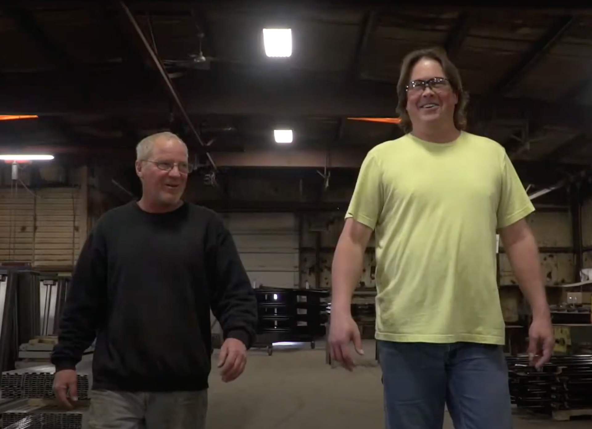 Two men are walking in a warehouse and one is wearing a yellow shirt.