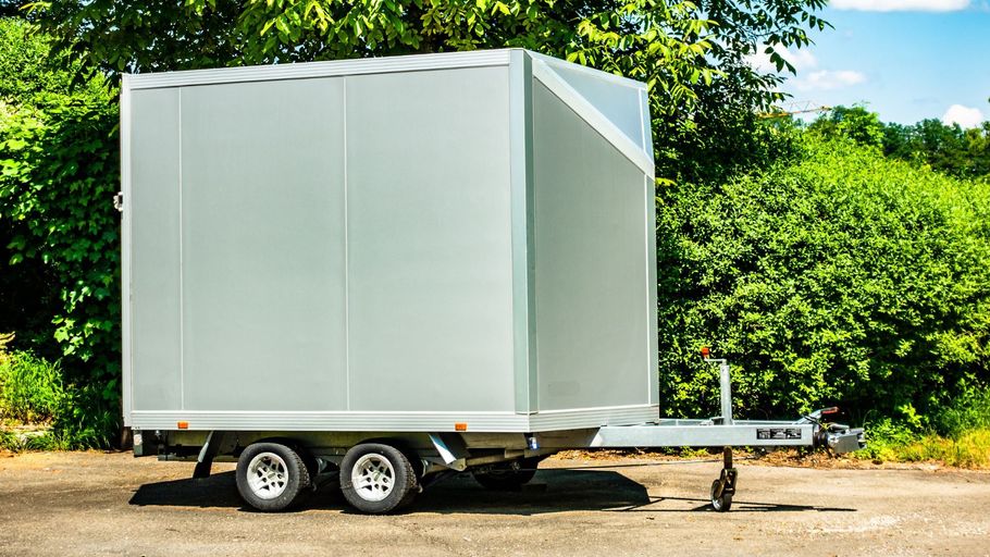 White enclosed cargo trailer parked on a dirt path with green trees in the background